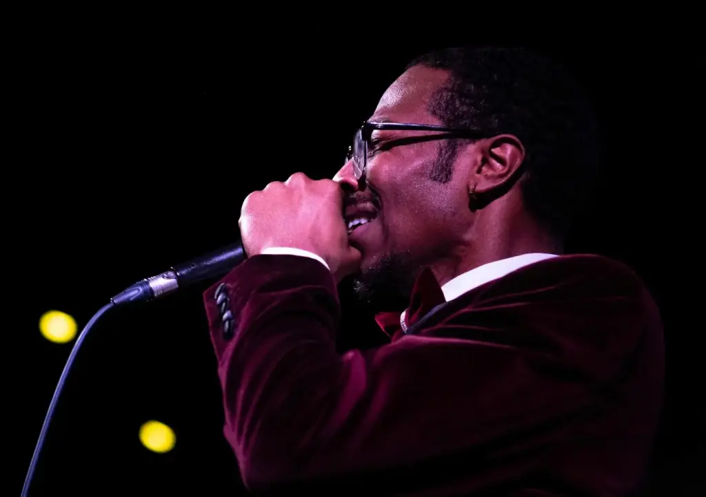 Traiveon singing (side profile, stage lighting, wearing glasses and maroon velvet blazer)