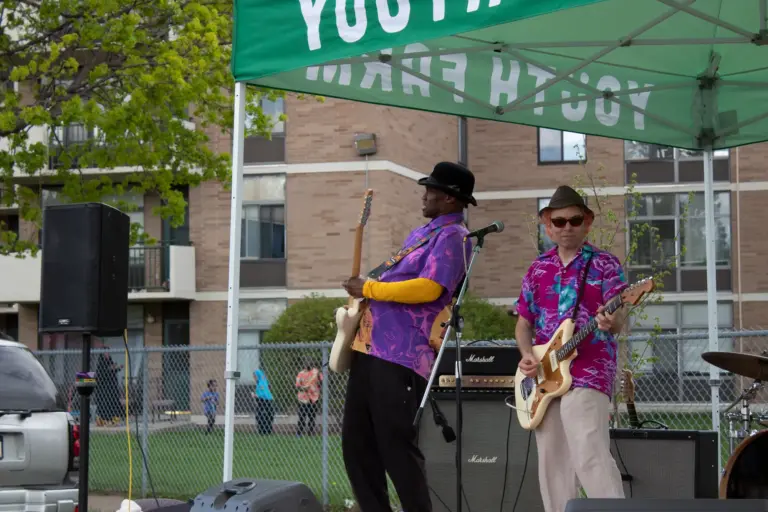 Jellybean Johnson playing an electric guitar at Imagine 6th Avenue North in 2022. Jellybean is a man with brown skin, wearing a purple short-sleeve button up shirt over an orange long sleeve undershirt. He is wearing a black fedora. Behind him, his bandmate, who has white skin, is also wearing a fedora and a printed pink button up short sleeve shirt. He is playing a white electric guitar.