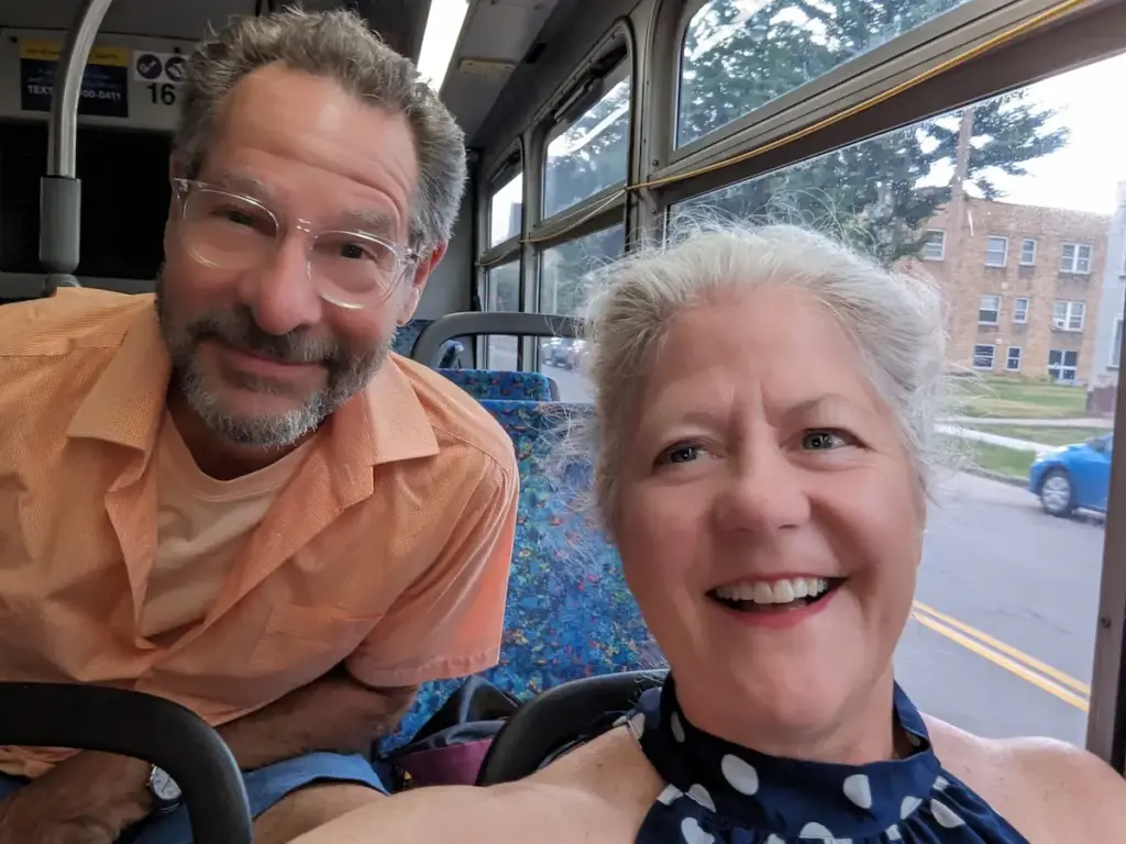 Selfie photo of Jim and Mary smiling while riding on a public bus. Jim is on the right in a light orange polo shirt and glasses with clear acetate frames. He is a white man with short grey hair and a neat beard and mustache. On the left is Mary, a white woman with light grey hair, pulled back from her face. She is wearing a blue sleeveless top.