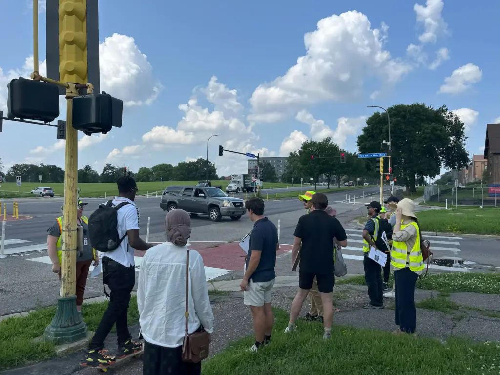 Person skateboarding on sidewalk while group of people in safety vests conduct safety audit walking tour of Olson Memorial Highway. It's a busy intersection, with a car taking a left turn